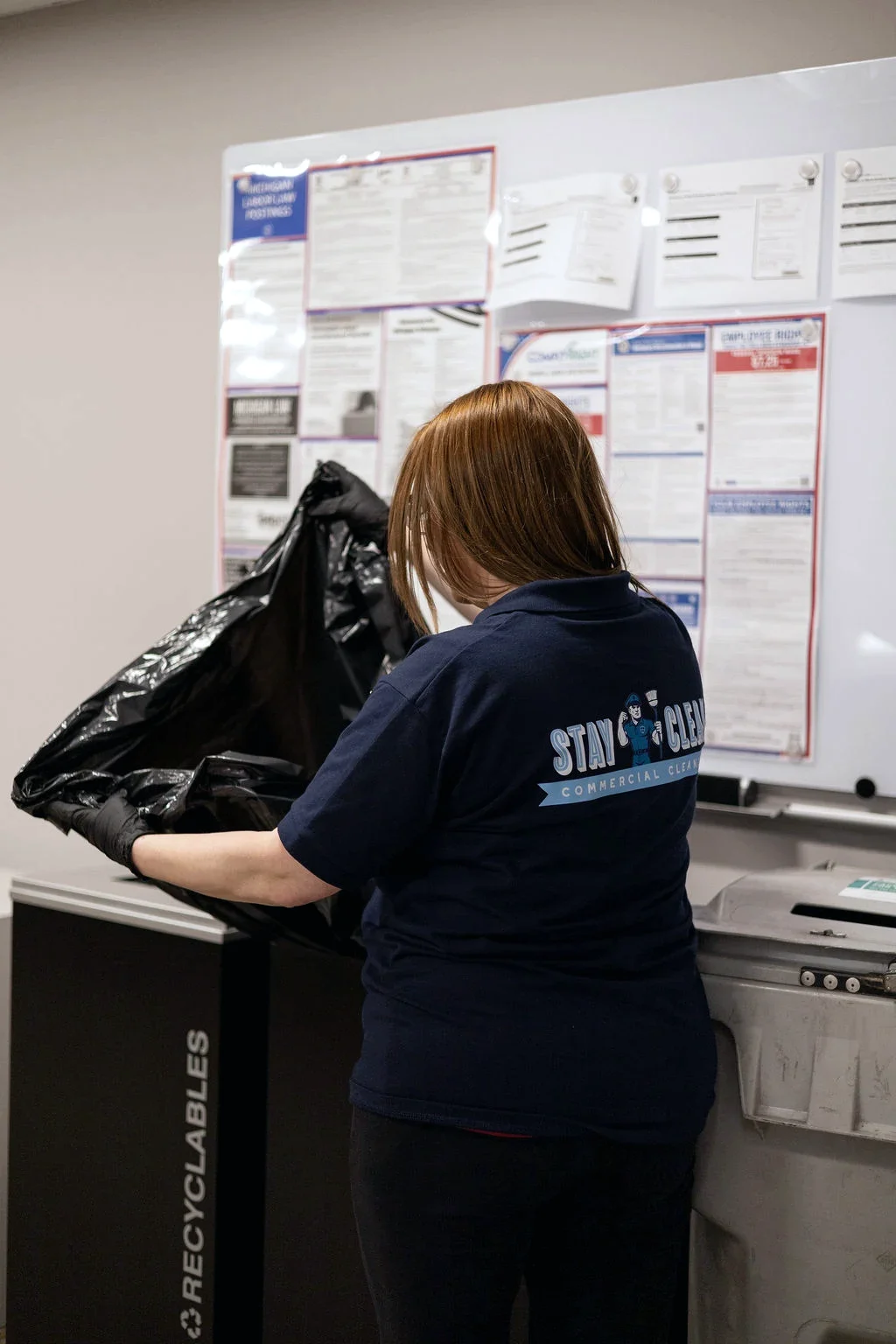 A Stay Clean team member handling waste and recycling beside a facility compliance board